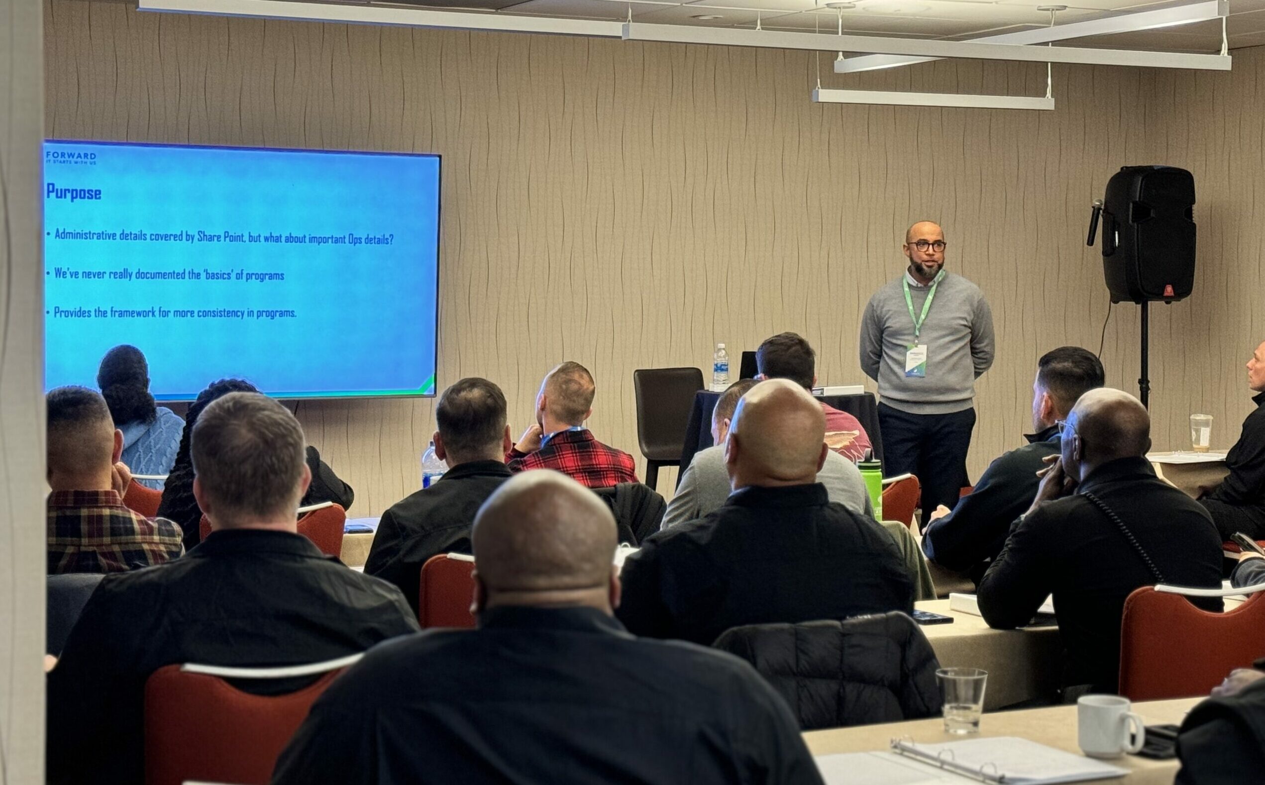 A man stands at the front of a room presenting a powerpoint to a room of people sitting at tables.